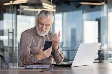 Modern bir ofiste, yaşlı bir işadamı akıllı telefon ve dizüstü bilgisayar kullanır, verileri analiz eder, video görüşmesi yapar veya profesyonel bir ortamda bilgi arar.