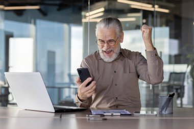 Gri saçlı yaşlı bir adam modern bir ofiste başarıyı kutluyor, akıllı telefonuna neşeyle bakıyor.