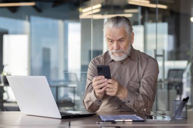 Yaşlı işadamı, profesyonellik ve odaklanma göstererek, modern bir ofiste akıllı telefonuna odaklandı.