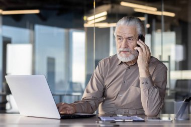 Üst düzey bir profesyonel aynı anda birden fazla iş yapıyor, telefonla konuşuyor ve çağdaş ofis ortamında dizüstü bilgisayar kullanıyor.