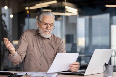 Kıdemli gri saçlı finansör mali raporları ve sözleşmeleri inceliyor, yetişkin patron sonuçlardan memnun değil ve hayal kırıklığına uğramış, kağıtlarla çalışıyor, iş yerinde laptop kullanıyor..