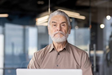 Modern bir ofiste, gri sakallı yaşlı bir adam profesyonel olarak odaklanmış ve kararlı bir dizüstü bilgisayar üzerinde çalışıyor.