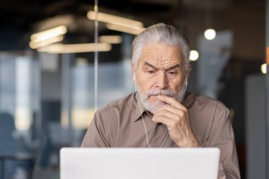 Yaşlı bir adam, modern ofis ortamında dizüstü bilgisayarı üzerinde düşünürken görülüyor. Bir göreve odaklanmış gibi görünüyor. Odaklanma ve tefekkür gösteriyor..