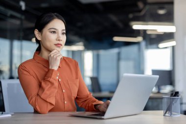 Derin düşüncelere dalmış, modern bir ofiste dizüstü bilgisayarla çalışan, odaklanma ve kararlılığı somutlaştıran kendine güvenen bir iş kadını..