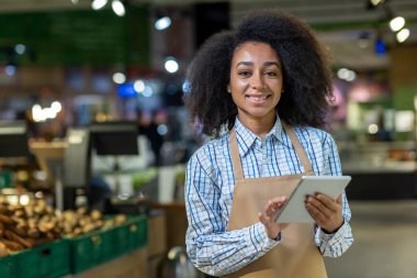 Modern bir süpermarkette çalışırken dijital tablet kullanan mutlu bir market çalışanı. Perakende ortamda neşe ve verimliliği yakalar.