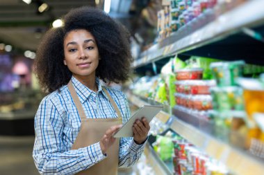 Elinde tabletle bir süpermarket çalışanı, koridordaki ürünleri işletiyor ve yönetiyor..