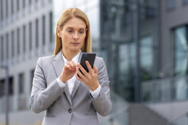 Gri takım elbiseli, modern bir ofis binasının önünde akıllı telefon kullanan kendine güvenen bir iş kadını. Ciddi ifade ile profesyonel ayarlar.