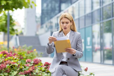 Takım elbiseli bir iş kadını, ofis binasının dışında sürpriz bir ifadeyle bir zarfı okuyor. Beklenmedik haber ve endişe kavramı.