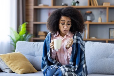 Battaniyeye sarılı hasta bir kadın, kanepede sıcak bir içecek tutarken termometreyle ateşini ölçüyor..