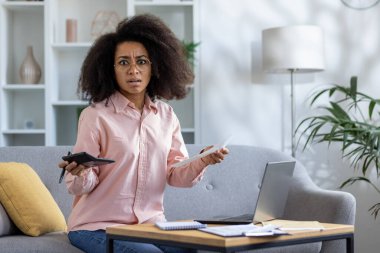 Stresli bir kadın oturma odasındaki bir kanepede otururken evinde evrak, hesap makinesi ve dizüstü bilgisayarla kişisel mali durumlarını yönetiyor..