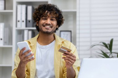 Elinde kredi kartı ve akıllı telefon tutan gülümseyen bir adam evde online alışveriş yapıyor. E-ticaret, alım, dijital ödeme ve modern yaşam tarzı kavramı.