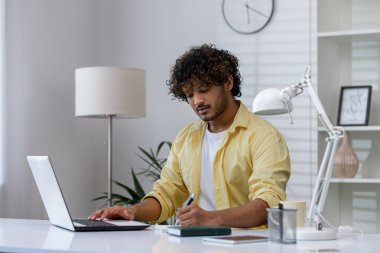 Sarı gömlekli bir adam evden uzaktan çalışıyor, dizüstü bilgisayar kullanıyor ve modern bir masadaki not defterine not alıyor..