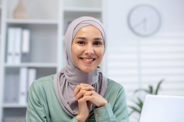 Kendine güvenen, tesettürlü, ofis masasında oturan, kendine güvenen, modern çalışma alanında çalışan bir kadın. İş ortamında profesyonellik ve pozitiflik.