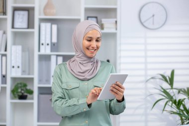 Tesettürlü gülümseyen kadın, modern ofiste kitaplıklar ve bitkilerle tablet üzerinde çalışıyor. Üretkenlik için teknolojiyi kullanan profesyonel iş kadını.