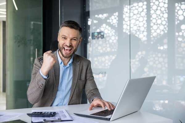 Возбужденный бизнесмен празднует успешное достижение с помощью кулачного насоса, сидя за столом в современной офисной среде.