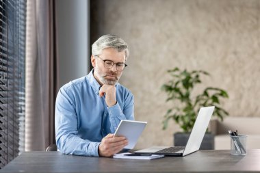 Üst düzey işadamı, büro masasında tablet üzerinde çalışıyor. Önemli görevleri düşünüyor. Laptop, bitki ve doğal ışık ile modern çalışma alanı.