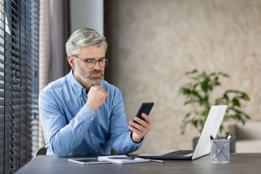Modern bir ofiste dizüstü bilgisayarla çalışırken akıllı telefona bakan olgun işadamı, odaklanmayı ve konsantrasyonu vurguladı.