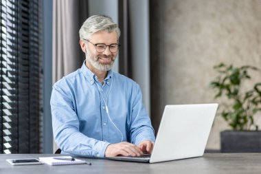 Gülümseyen olgun iş adamı bilgisayarını uzaktan veya çevrimiçi iletişim için kullanıyor. Ev ya da ofis ortamından profesyonel çalışma.