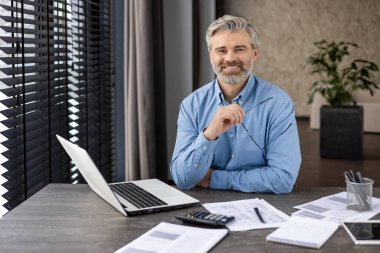 Kendine güveni tam bir işadamı. Masasında dizüstü bilgisayar ve belgelerle çalışırken gülümsüyor. Modern bir ofis ortamında..