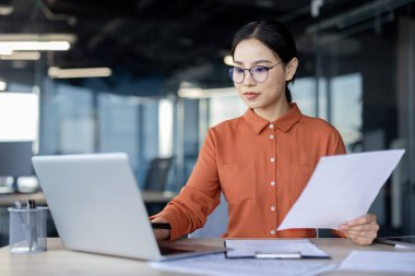 Ofis içinde belgelerle çalışan Asyalı finans kadını, evrakların arkasındaki kağıtlar, iş yerinde mali raporlara bakan ciddi iş kadını, kendine güvenen başarılı kadın muhasebeci..