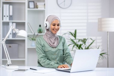 Tesettürlü, kulaklık takan, modern bir ofiste dizüstü bilgisayarla çalışan gülümseyen bir kadın. Bitkiler ve beyaz dekorlu profesyonel çalışma alanı.