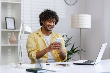 Gülümseyen adam ofis masasında bilgisayarını ve modern dekorunu kullanarak bir anın tadını çıkarıyor..