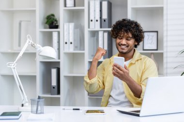 Modern ofis ortamında akıllı telefonuna bakarken başarıyı kutlayan mutlu bir adam. Neşeli ifade başarıyı ve pozitifliği ifade eder..