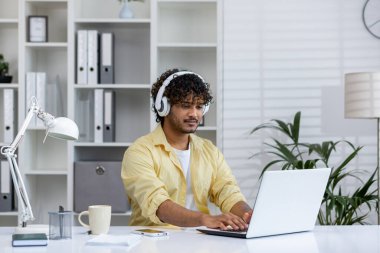 Evden çalışan genç bir profesyonel, kulaklık takıyor ve modern bir ofis alanında dizüstü bilgisayar kullanıyor. Uzak çalışma ve üretkenlik kavramı.