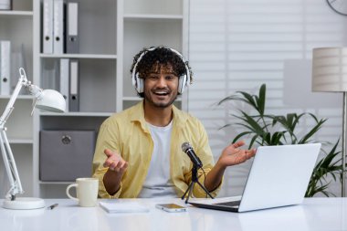 Gülümseyen adam, kulaklıklar, mikrofon ve dizüstü bilgisayarla ev sahipliği yapıyor. Neşeli ve ilgi çekici ses içeriği yaratıyor..