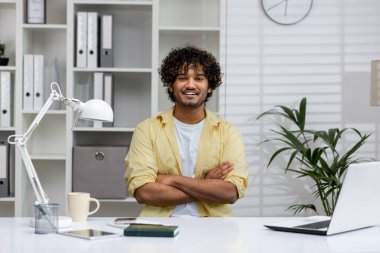 Kıvırcık saçlı gülümseyen adam, kollarını kavuşturmuş bir şekilde ofis masasında oturuyor. Modern bir çalışma alanında kendinden emin ve rahat görünüyor..