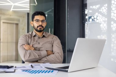 Kendine güveni tam bir iş adamı elinde dizüstü bilgisayar ve belgelerle modern ofis ortamında profesyonel ve odaklanmış bir tavır sergiliyor..