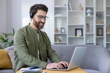 Parlak, modern bir oturma odasında dizüstü bilgisayar ve kulaklık kullanarak evden çalışan gülümseyen adam..
