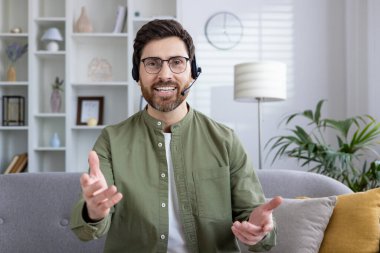 Kulaklığı olan gülümseyen bir adam evinde otururken video görüşmesi yapıyor. Uzak çalışma ve sanal iletişim kavramı.