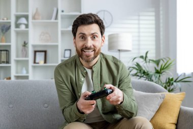 Evde video oyunu oynayan, rahat bir koltukta oturan, modern bir oturma odasında boş vakitlerin tadını çıkaran bir adam..