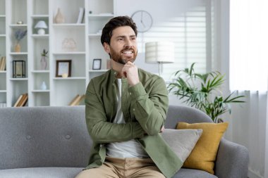 Gri bir koltukta oturan sakallı adam şık bir oturma odasında, düşüncelere dalmış ve gülümsüyor. Bitkiler ve dekorlarla dolu, parlak ve rahat bir ev..