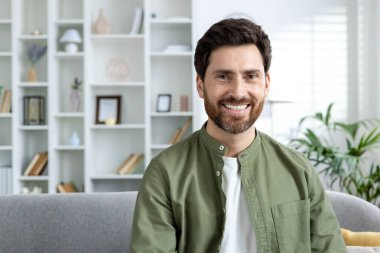 Oturma odasındaki kanepede oturan gülümseyen bir adamın portresi, arka planda raflar ve bitkiler var..