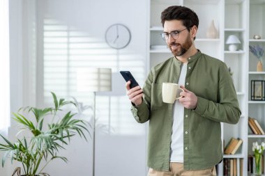 Elinde kahve fincanı tutan sıradan bir adam ve parlak bir evde akıllı telefon kullanıyor. Rahat atmosfer, modern yaşam tarzı.