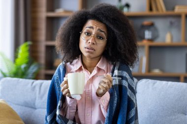 Kadın kendini kötü hissediyor, battaniyeyle kaplı bir kanepede otururken elinde bardak ve termometre tutuyor. Hastalık, iyileşme ve kişisel bakım kavramı.