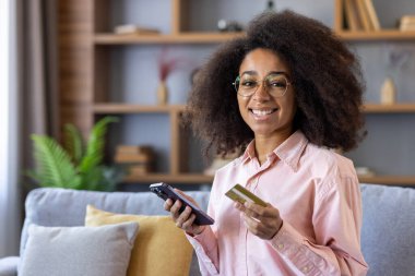 Mutlu kadın evde rahat bir koltukta otururken online alışveriş için akıllı telefon ve kredi kartı kullanıyor..