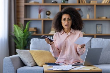 Şok olmuş bir kadın elinde telefonla faturaya bakıyor. Evdeki bir kanepede oturuyor..