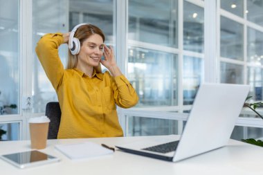 Modern, parlak bir ofis ortamında dizüstü bilgisayarla çalışırken kulaklıkla müzik dinleyen bir kadın. Rahatlamış ve mutlu görünüyor..