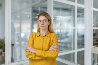 Kendine güvenen, sarı tişörtlü, kolları çapraz modern bir ofiste duran bir iş kadını. Çağdaş bir iş yerinde profesyonel ve kararlı bir kadın lider..