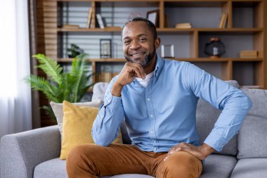 Gülümseyen adam şık ve modern bir oturma odasındaki koltukta rahatça oturuyor. Doğal ışık alanı aydınlatır..