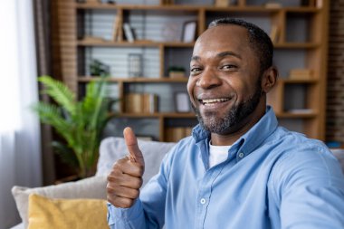 Mavi gömlekli mutlu bir adam, rahat bir evde video görüşmesinde başparmak işareti yapıyor..