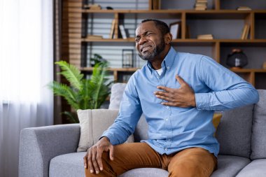 Göğsünü tutan, koltukta rahat bir şekilde otururken acı çeken kalp krizi ya da göğüs ağrısı belirtileri gösteren bir adam.