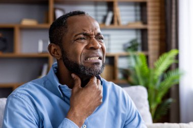 Evde oturan, boğazında rahatsızlık ya da ağrı hisseden ve boynuna dokunan bir adam. Görüntü hastalık, rahatsızlık ya da sağlık durumunu aktarır..