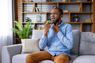 Adamın boğazında ağrı ve boğaz spreyi var. Rahat bir evde, kanepede otururken, sıkıntılı görünüyor.