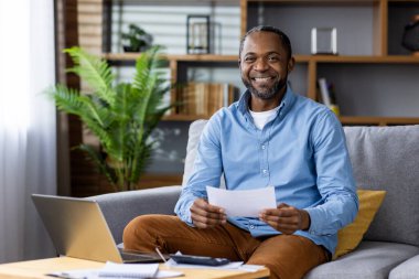 Mavi tişörtlü neşeli bir adam kanepede oturuyor ve elinde bir dizüstü bilgisayarla çalışıyor..