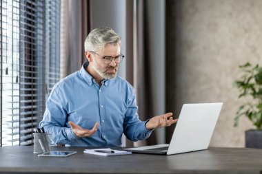 Üst düzey işadamı, büyük pencereleri ve fabrikası olan modern bir ofiste dizüstü bilgisayarla yapılan bir video görüşmesi sırasında hayal kırıklığını dile getiriyor..
