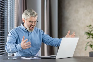 Mavi gömlekli kıdemli işadamı, modern ofis ortamında dizüstü bilgisayarla çalışırken başarıyı kutluyor..